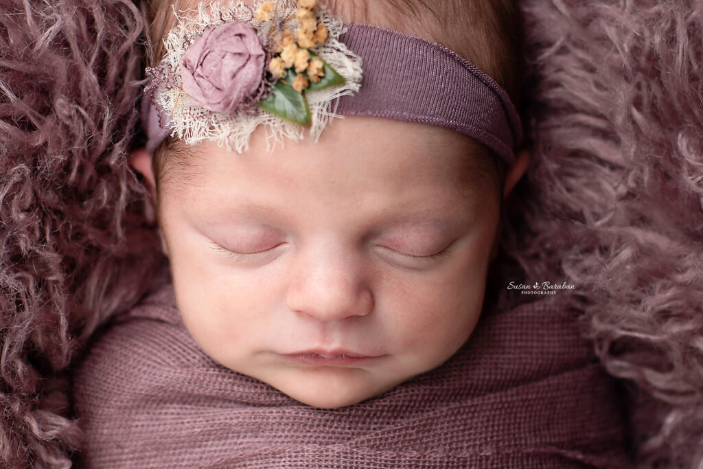 Close up of a sleeping newborn baby girl wearing a purple swaddle, purple headband and laying in a basket with a purple fur.