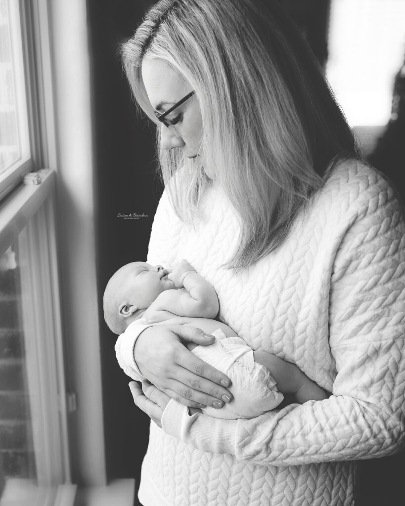 Black & white image of a mother standing near a window gazing down at her newborn baby girl sleeping in her arms during a Frisco newborn photo session.