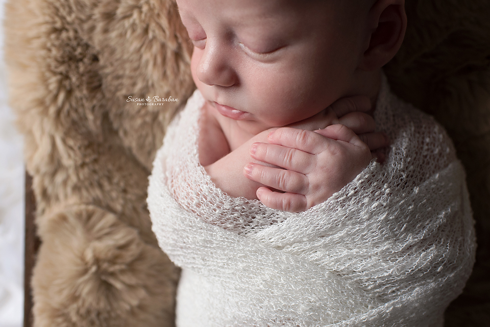 Close up image of a newborn boy sleeping with his hands crossed while wearing a white swaddle.
