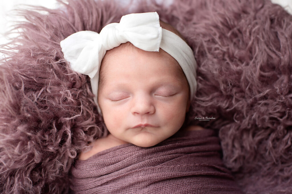 Newborn girl wearing a large white headband with a bow and wrapped in a purple swaddle during her newborn session with Allen, TX newborn photographer Susan Baraban Photography.
