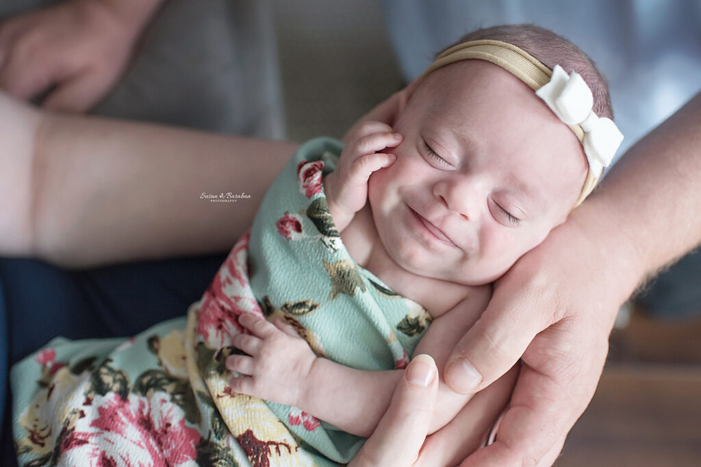 Newborn baby girl wrapped in a floral swaddle, smiling while in her parent's arms.