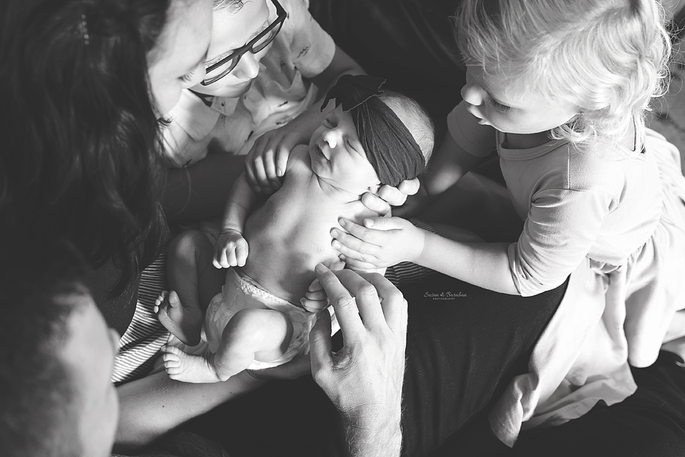 Black and white image of an adorable family holding a newborn baby girl who is wearing a headband with a bow during their lifestyle newborn session in Prosper, TX.