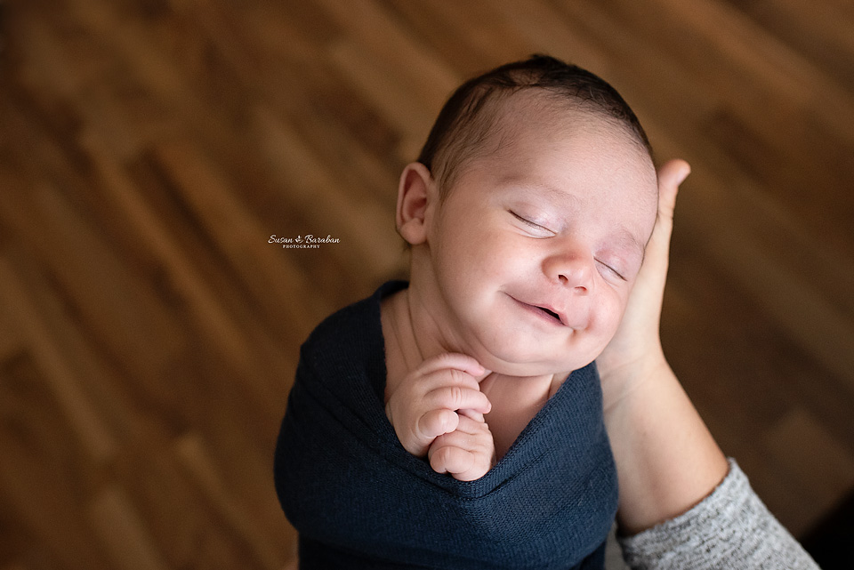 Newborn boy wrapped in a navy blue swaddle and smiling while his mother holds him at his in-home newborn photo shoot.