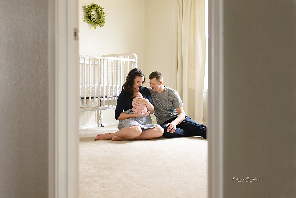 New parents sitting on the floor of the newborn nursery holding their baby girl wrapped in a pink swaddle during her lifestyle newborn photo session at their home in Prosper.