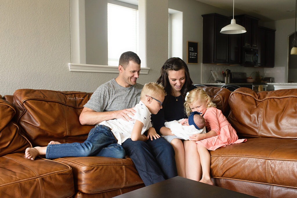 Family of five sitting on the couch in their Prosper, TX home admiring their newborn baby girl at her newborn photo shoot with Susan Baraban Photography.