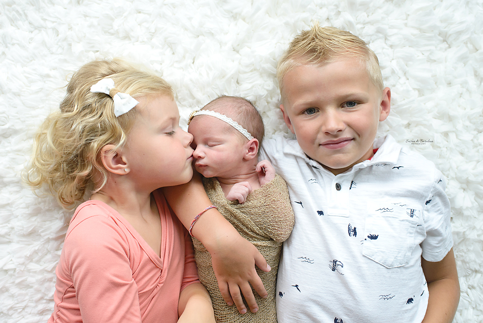 Two older siblings have their arms around their newborn baby sister.  The older sister is kissing her nose at their Prosper Newborn Photo Shoot.