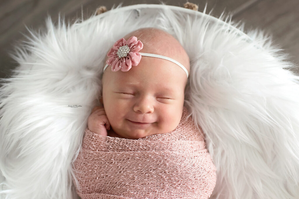 Newborn girl in a pink wrap wearing a pink flower headband smiling while laying in a white basket with a white fur.