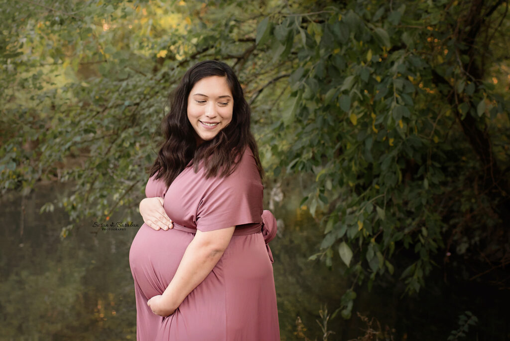 Pregnant woman cradling her belly while standing in front of trees and a small pond at Prairie Creek Park in Richardson, TX.