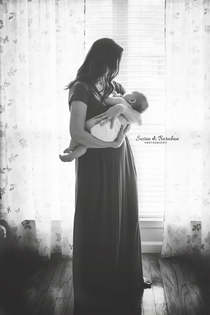 Mother cradling her newborn baby while standing in beautiful window light at home during a newborn photo shoot.
