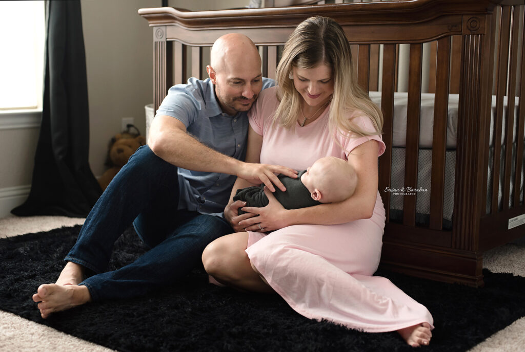 New parents holding their son while sitting in front of a crib in their son's nursery during his lifestyle newborn photo session with Susan Baraban Photography.