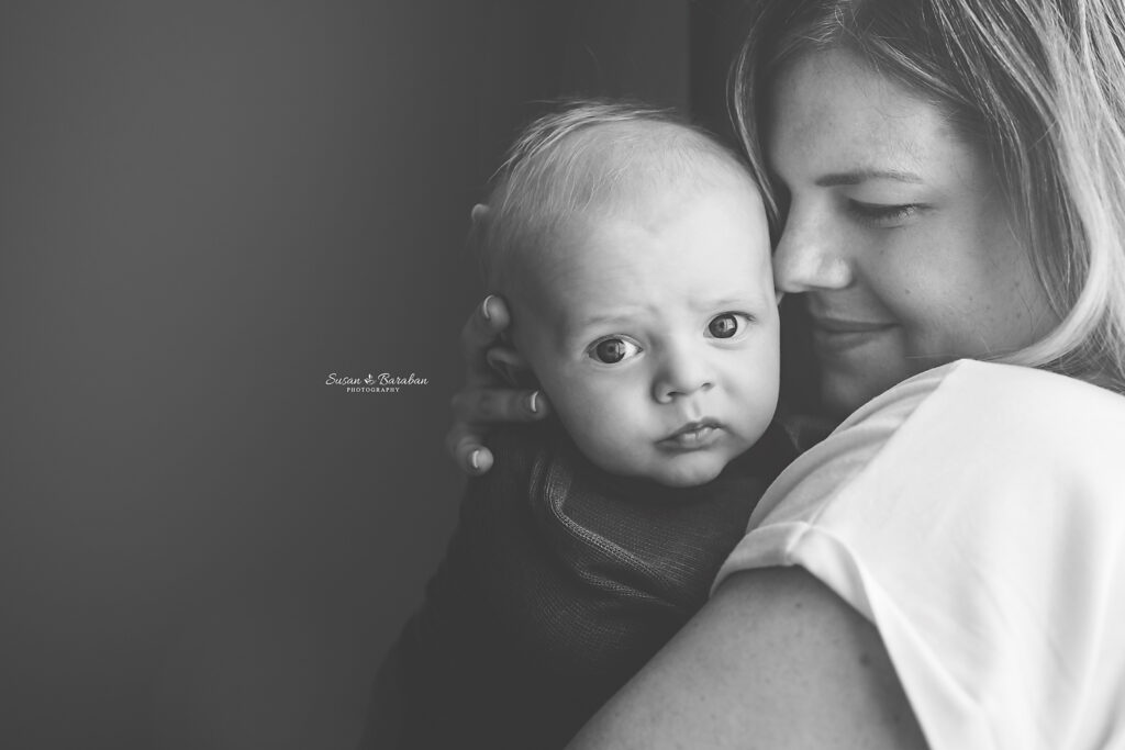 Black & White image of a 3 month old baby looking directly in the camera while his mother snuggles him.