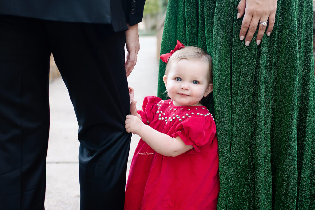 Adorable toddler in a red dress holding on to her father's leg and smiling at the camera.