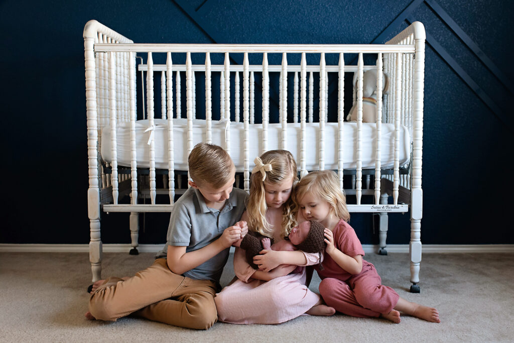 Siblings cuddling a newborn baby boy while sitting on the floor of his nursery.  Wall painted dark blue, white crib.