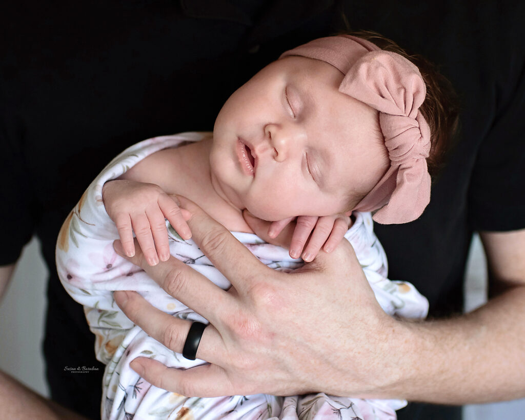 Newborn baby girl wrapped in a floral swaddle and matching pink bow headband sleeping in her father's arms.