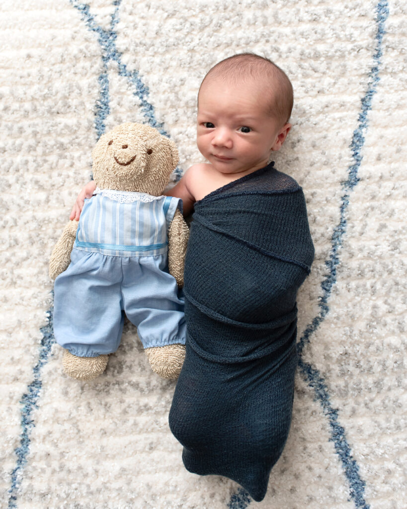 Newborn boy wrapped in a navy blue swaddle cuddling with a sentimental small teddy bear at his newborn photo session in Dallas, TX.